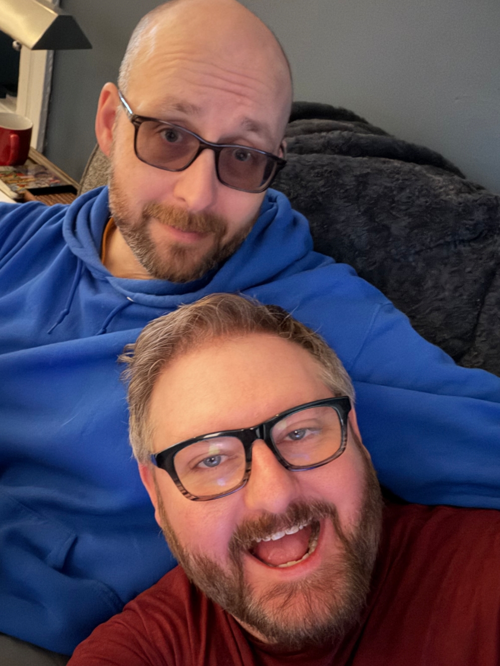 Me and my husband smiling both with glasses relaxing together on a couch, my husband in a blue hoodie leaning back and myself in a red shirt in the foreground.